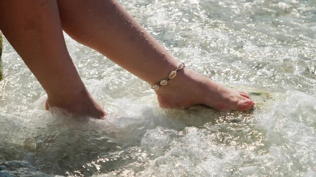 Extreme close-up of a shell and blue bead anklet on a bare foot on a wet rock, wave splashing in background, handheld shot.