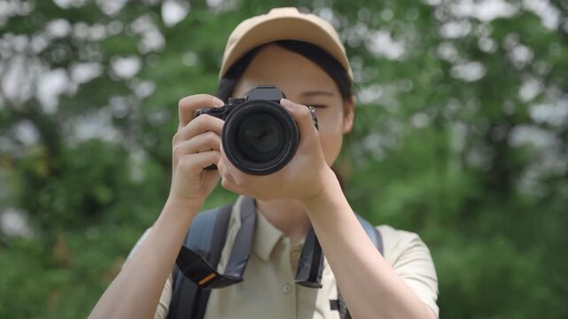 Woman photographer with big backpack wearing trekking clothes raises DSLR digital camera to her face and takes a photo of beautiful nature forest landscapes, travel adventure, outdoor exploration