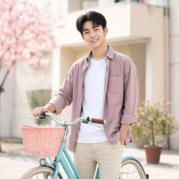 Young man stands smiling with a pastel bicycle and pink basket on a tree-lined residential street in spring. Casual outfit and relaxed pose convey warmth and outdoor leisure.