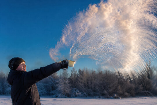 Man throws hot water into freezing air creating a dramatic steam arc at sunrise. Scene shows winter landscape with frosty trees and clear blue sky.
