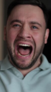 Vertical video Close up of a young stressed man shouting on living room background. Motions concept. Man's reaction to bad news.