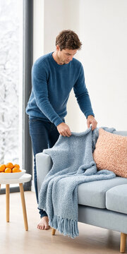 Man arranges a knitted blanket over a sofa cushion in a bright living room, suggesting home comfort and interior styling.