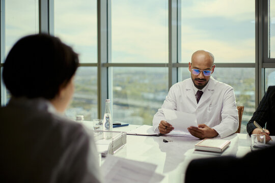 Medium shot of middle aged male doctor reviewing paperwork during meeting with colleagues, discussing medical administration and leadership. Ideal for healthcare management or policy marketing