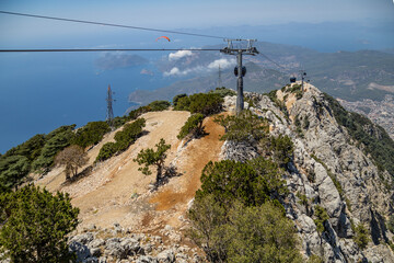 Cable car gondola lift on steel cables over rocky mountain ridge with paraglider in sky. Aerial sea bay panoramic view, sunny summer daytime, Babadag, Oludeniz, Fethiye, Turkey. © Vlad Rakin