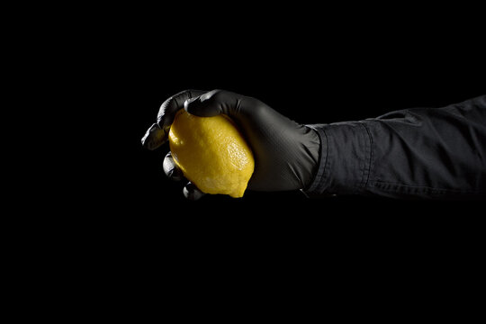 Hand in black glove ruthlessly squeezing a lemon on black background for corporate audit and debt concept with copyspace