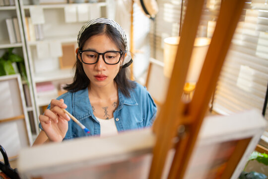 Gen Z Asian woman freelance artist painting with paintbrush on canvas easel, representing creative professional women designer, online independent creator and art studio hobby lifestyle