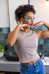 African American woman standing at home vanity wearing tank, posing with blue eye patches