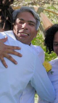 Vertical video: Smiling grooms hugging as guest approaching from right cheering at garden arch