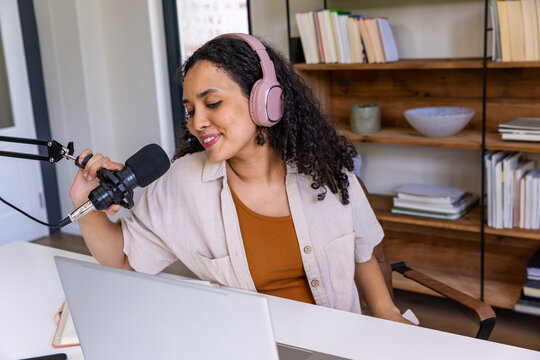 African American woman recording at home desk speaking into microphone with pink headphones