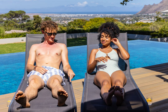 Diverse couple reclining on deck loungers by pool edge, wearing swimwear and sipping orange drinks