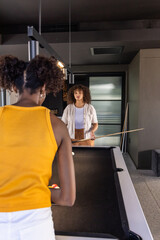 Naklejka premium Diverse female friends wearing yellow tank playing pool in game room with cues, copy space