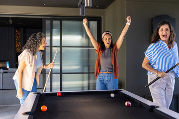 Naklejka premium Three female friends playing in modern game room, celebrating at pool table with cues and balls
