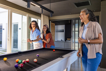 Naklejka premium Diverse female friends holding cues and playing pool around black-felt table at home, laughing