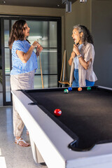 Fototapeta premium Diverse female friends standing holding wood cues talking by black pool table in home game room