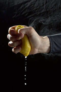 Male hand forcefully squeezing lemon with dripping juice and smoke on dark background margin squeeze