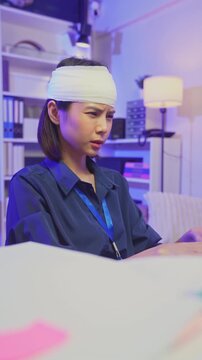 Injured young Asian office worker wearing head bandage while working on laptop in office, health and work pressure concept.