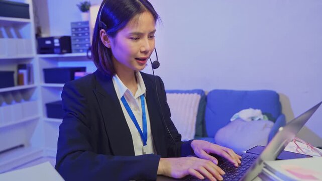 Young Asian customer service representative wearing microphone headset and working on laptop in modern office.