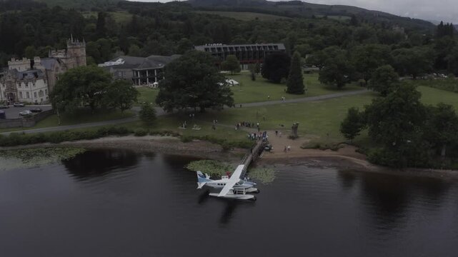 High angle aerial shot of seaplane docked at wooden pier on loch lomond scotland with historic castle hotel and lush green hills in background during overcast summer day with tourists on shoreline.