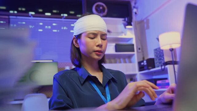 Injured young Asian office worker wearing head bandage while working on laptop in office, health and work pressure concept.