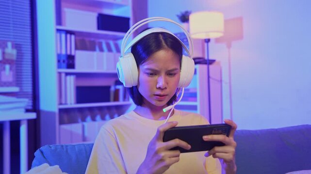Excited young Asian girl playing mobile video game with headset at home.