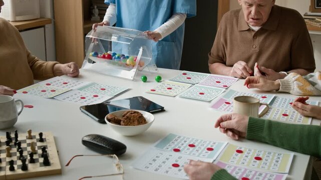 Close up shot of seniors playing bingo game with female nurse spinning lottery drum in retirement home. Clip for promoting geriatric healthcare and assisted living facility services
