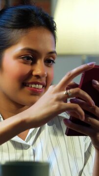Indian businesswoman checking mobile phone at desk in modern cozy office, Asian female glued to smartphone while multitasking during office hours, smiling with positive body language in workplace