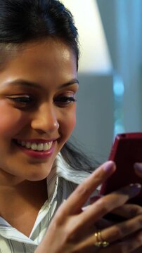 Indian businesswoman checking mobile phone at desk in modern cozy office, Asian female glued to smartphone while multitasking during office hours, smiling with positive body language in workplace