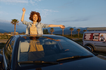 Obraz premium Joyful woman with curly hair stands through a sunroof of a car, arms raised against a bright sky and palm silhouettes, embodying lifestyle, travel, and freedom.