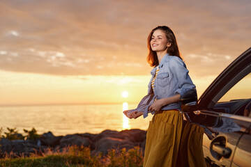 Fototapeta premium Smiling woman leaning against a car at sunset, enjoying a relaxed lifestyle in casual denim and a jacket, with golden hour light and warm outdoor vibes.