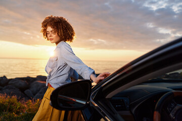 Fototapeta premium Young woman with curly hair steps away from a dark car at sunset along a rocky coast, wearing a light denim shirt and mustard skirt, looking contemplative.