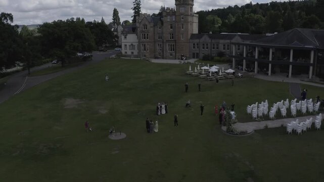 Birds eye view cinematic footage captures distant wedding party guests gathered on green grass lawn in front of historic scottish castle and modern hotel building on an overcast day.