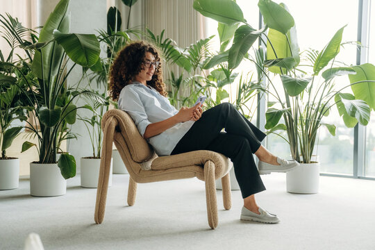 Young woman with curly hair sitting on a modern chair using smartphone surrounded by large indoor plants in a bright, airy room with natural light