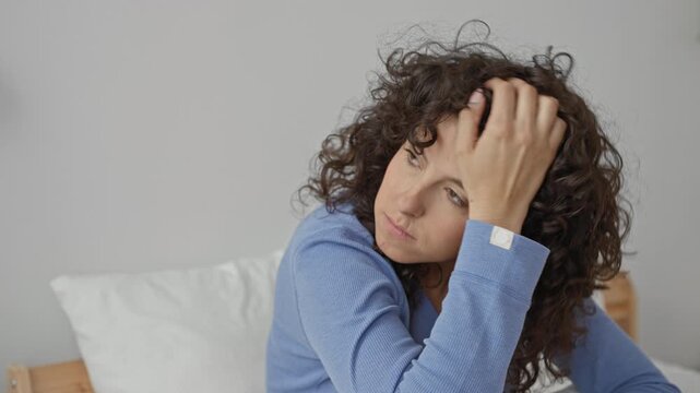 Woman on bed holding head with hand in building, curly hair visible, eyes closed leaning forward touching forehead and temple; fatigue reflection.