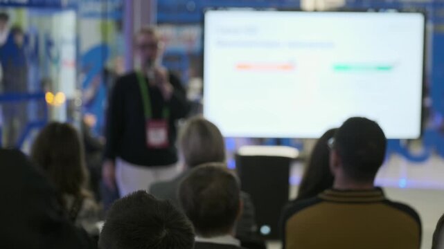 Audience listens to presenter during corporate conference workshop. Blurred background, projection screen, rows of attendees, focused presentation atmosphere.
