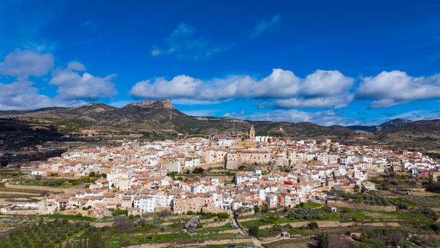Wide panoramic view of Chelva, inland Valencia, Spain, with baroque church tower and blue dome at centre, terraced orchards in foreground and limestone mountains under dramatic cloudy sky