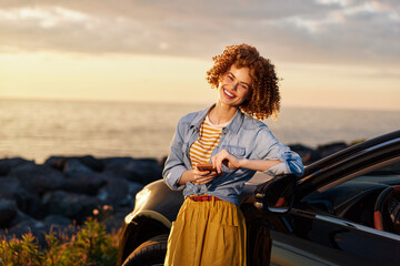Fototapeta premium Smiling woman with curly hair leaning beside a car at sunset, wearing a denim shirt and mustard skirt. Outdoor lifestyle, travel vibe, carefree mood, warm colors, golden hour lighting.