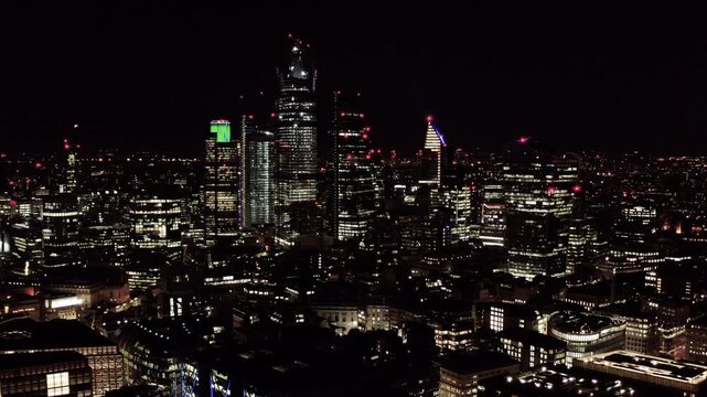 Aerial drone night view over the City of London financial district, England, showing illuminated skyscrapers including 22 Bishopsgate and Heron Tower rising above surrounding office buildings.