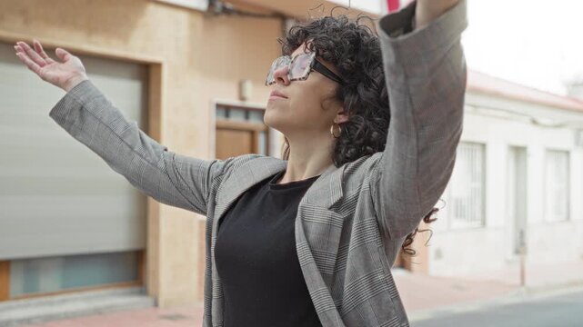 Woman with glasses wearing a plaid blazer and black knit top raises arms and tilts head while looking upward on street; serenity calm gratitude.