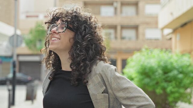 Young woman smiles and looks up wearing oversized tortoiseshell glasses on a street by an apartment building; confidence curiosity.