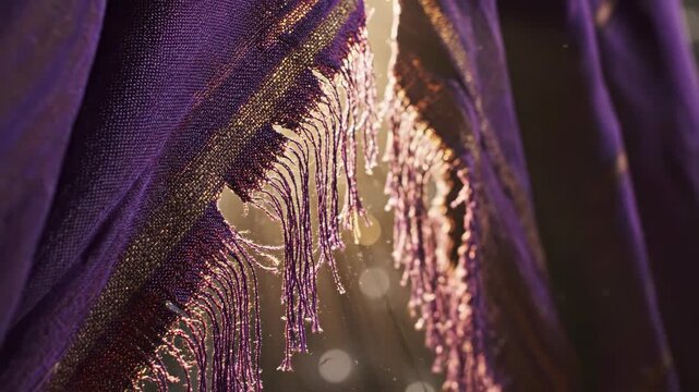 Close-up of purple fabric with gold threads and frayed fringe. Backlit textile with floating dust particles in warm sunlight. Luxury material detail