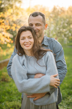 Happy romantic couple portrait. Sincere middle aged husband and wife embrace closely with heads together, smiling warmly in sunny spring or autumn garden at sunset, sharing a tender moment of love.