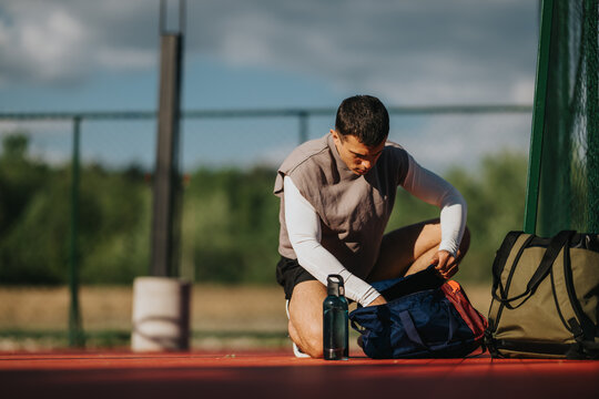 A young male athlete kneels and packs his sports bag beside a water bottle on an outdoor court. He prepares gear and equipment before a training session on a fenced sports court.