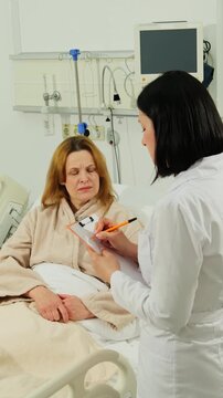 Doctor records fresh notes while woman patient rests in hospital bed. Medical specialist writes followup remarks reviewing patient status