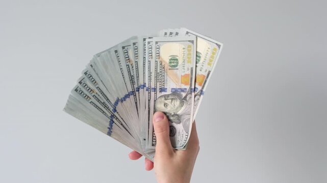 Hand holds fanned bills against plain background. Fingers grip stack of cash, showing wealth clearly. Neutral expression, no visible emotion on face. Perfect for finance, business