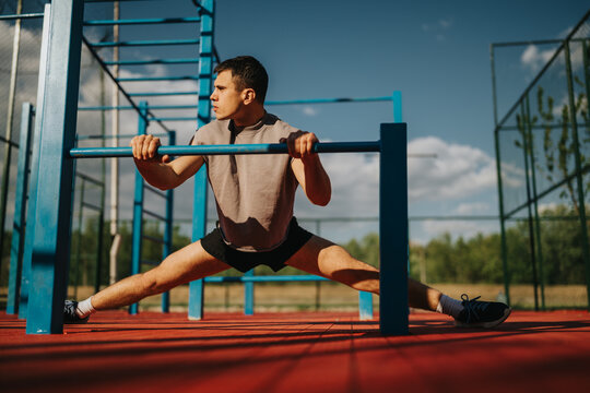 Young man performs a wide straddle stretch while holding onto outdoor calisthenics bars. An athlete stretches across a park exercise area during a training session.