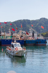 Fototapeta premium Dalian, China-June 5, 2026: Small motorboat moored in calm harbor with fishing vessels and China flags against green hills at Fisherman’s Wharf is one of the city’s most photogenic seaside spots