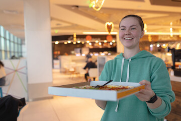 Fototapeta premium Woman presents open pizza box forward with cheerful smile, warm decorative lights create festive mood, casual hoodie, indoor cafeteria environment, soft blur emphasizes human expression.