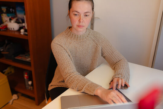 Close view indoor scene with woman typing laptop, textured sweater details, concentrated facial expression, neutral tones, warm home office setup, natural soft light.