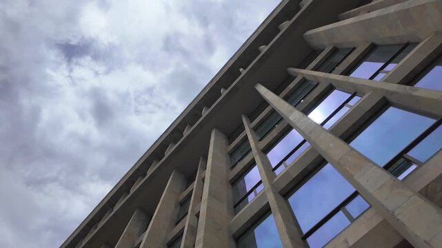 Low angle view of a modern brutalist building with a concrete facade and large windows reflecting the cloudy sky, capturing a sense of imposing architectural geometry and urban design