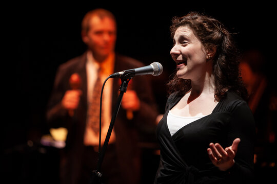 Jazz Singer. A woman singing into a stand microphone on stage with a blurred background figure.